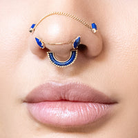 A close-up of a person's nose wearing a gold nose ring with blue stone accents, showing the jewelry on the nasal bridge and sides of the nose.